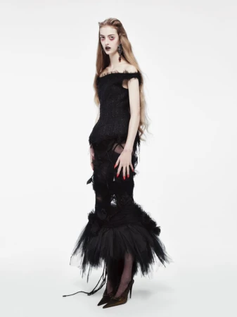 Model with long blonde hair in a black off-the-shoulder dress standing against a seamless white studio background