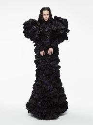 Model standing against a plain white background wearing an elaborate, heavily textured black ruffled gown with exaggerated sculptural shoulders.