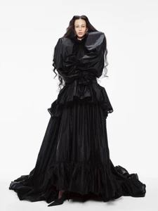 Model wearing a voluminous black gown with exaggerated shoulders standing against a white seamless background