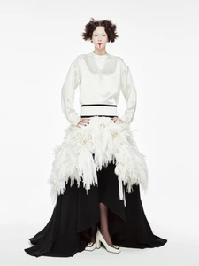 Model standing against a white seamless background wearing a white top and a feathered skirt over a black gown