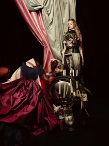 Model standing before draped fabrics with a round mirror, candles, and a burgundy skirt in a dark studio setting.