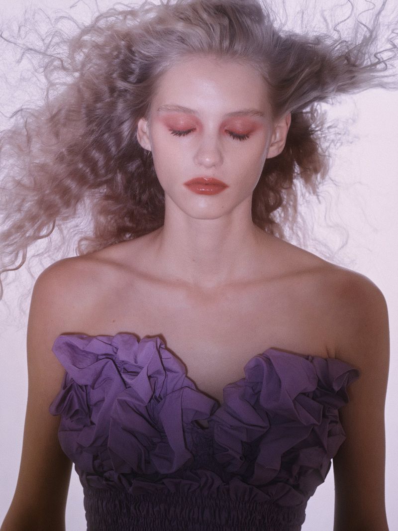 Close-up portrait of a fair-skinned woman with voluminous curly hair, wearing a purple strapless dress, with pink makeup and closed eyes.