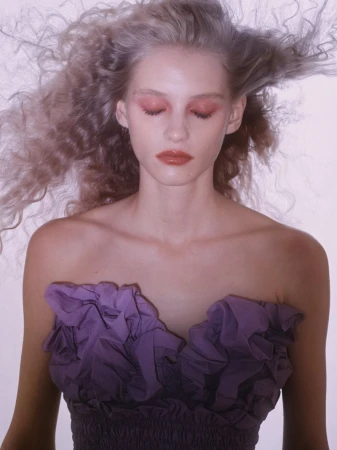 Close-up portrait of a fair-skinned woman with voluminous curly hair, wearing a purple strapless dress, with pink makeup and closed eyes.