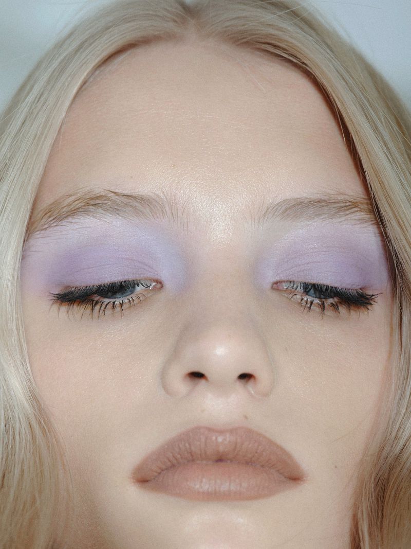 Close-up of a woman's face with platinum blonde hair, featuring purple eyeshadow, nude lipstick, and dramatic eyelashes.