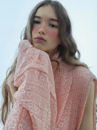 A young fair-skinned woman with wavy brown hair poses indoors wearing a peach patterned dress, looking directly at the camera.
