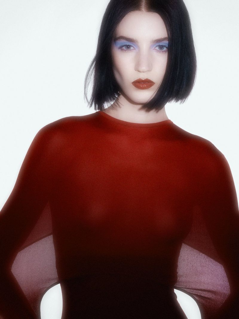 Portrait of an adult woman with short, dark hair wearing a maroon top and blue eyeshadow against a bright white background.