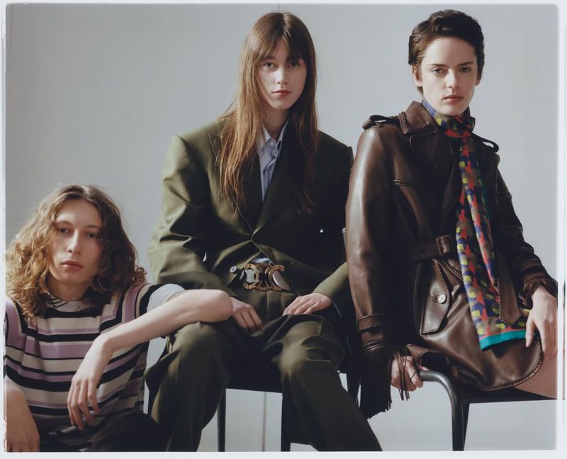 Three fashion models sit in a photo studio. Two women and one man model high-fashion clothing in a brightly lit studio setting.