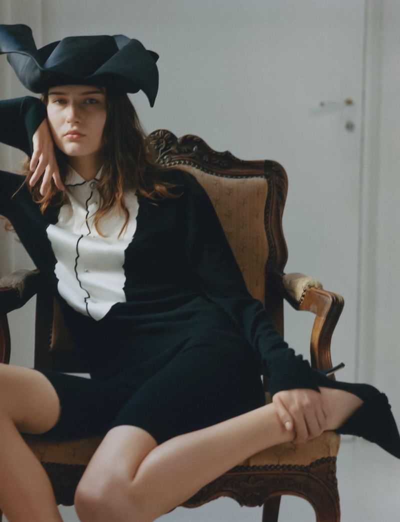 A young woman wearing a large, structured black hat and a black sweater with a white collared bib sits in a carved wooden armchair.