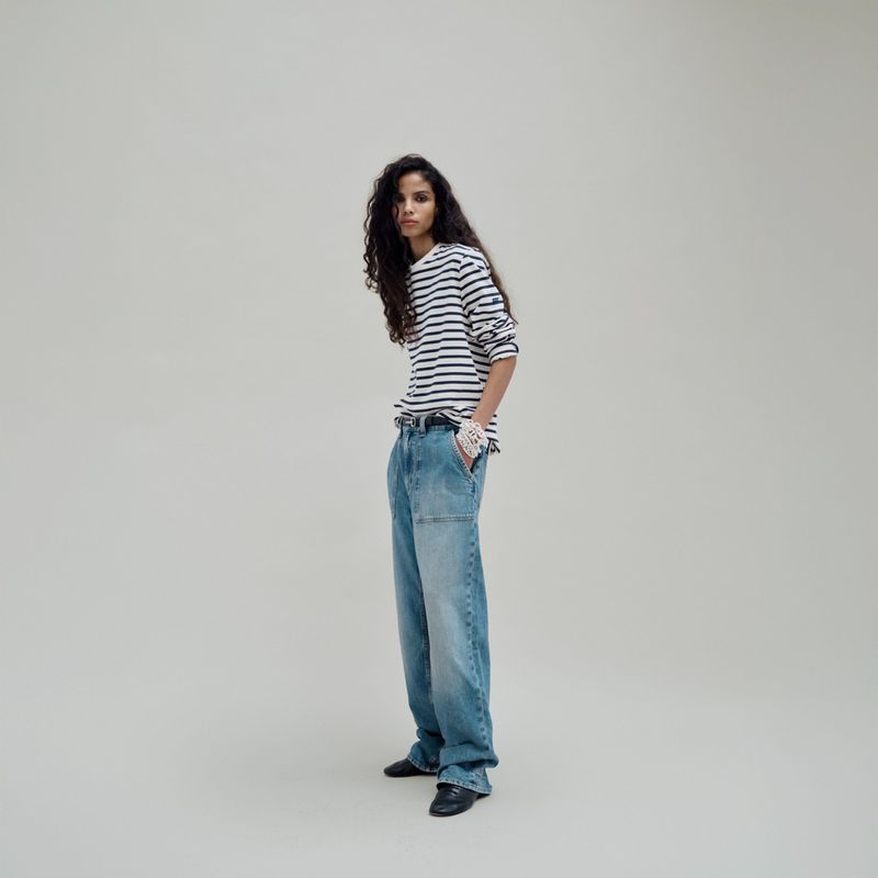A slender adult fashion model with long dark hair poses in a studio wearing a striped top and baggy jeans against a neutral backdrop.