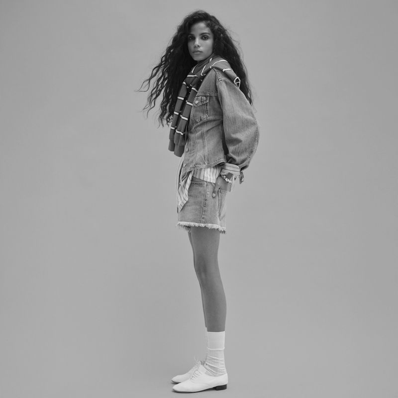 Black and white portrait of a woman wearing a denim jacket and shorts, white socks, and white shoes against a light grey background.