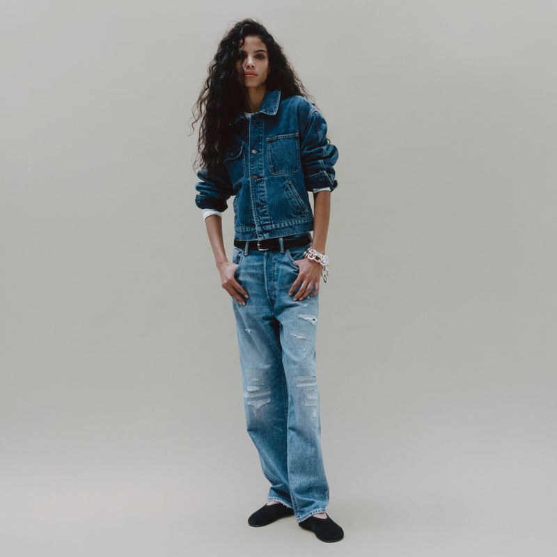 Model with long dark wavy hair wearing a denim jacket and blue jeans against a neutral grey studio backdrop.