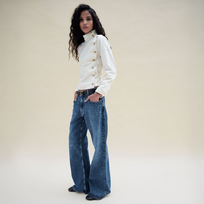 Model with long dark wavy hair wearing a white buttoned high-neck top and blue denim jeans against a seamless neutral background