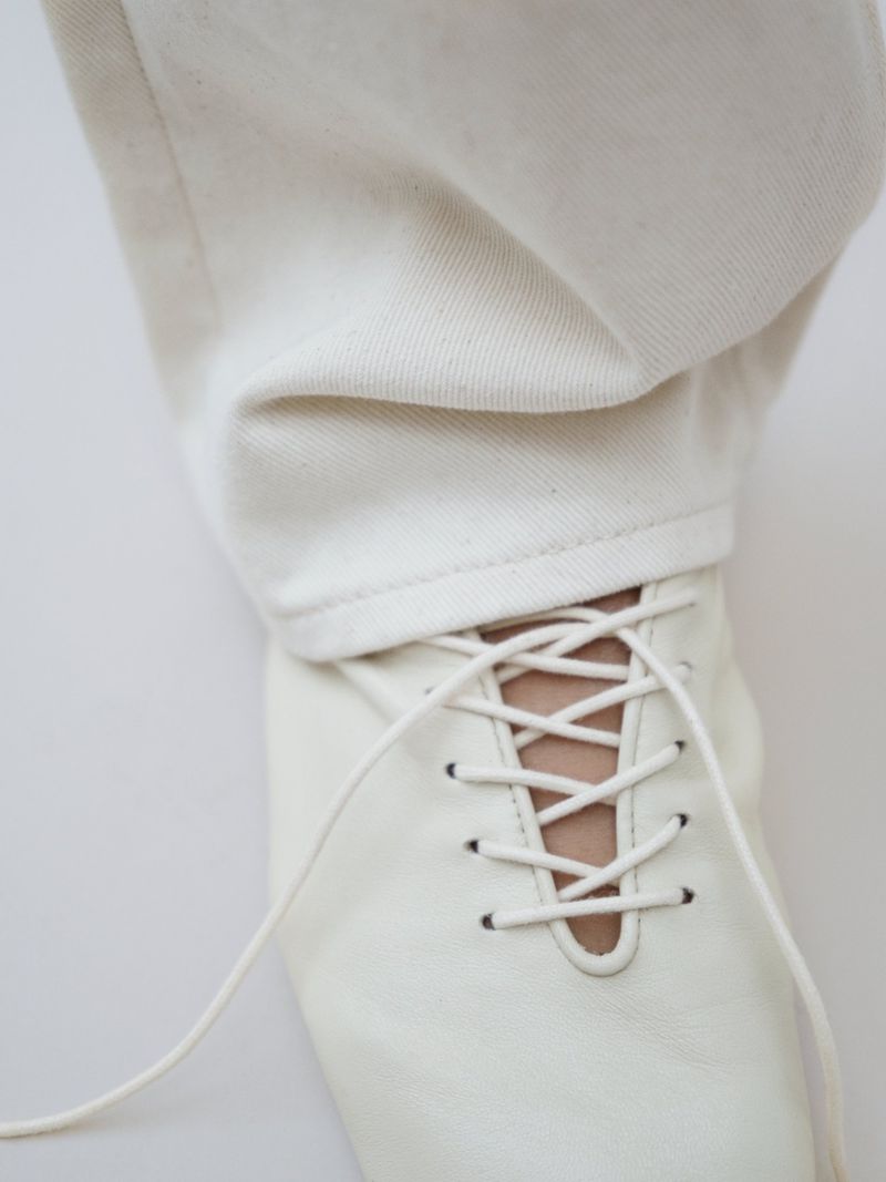 A close-up of a person wearing white denim jeans and white lace-up shoes against a plain white background in a studio setting.