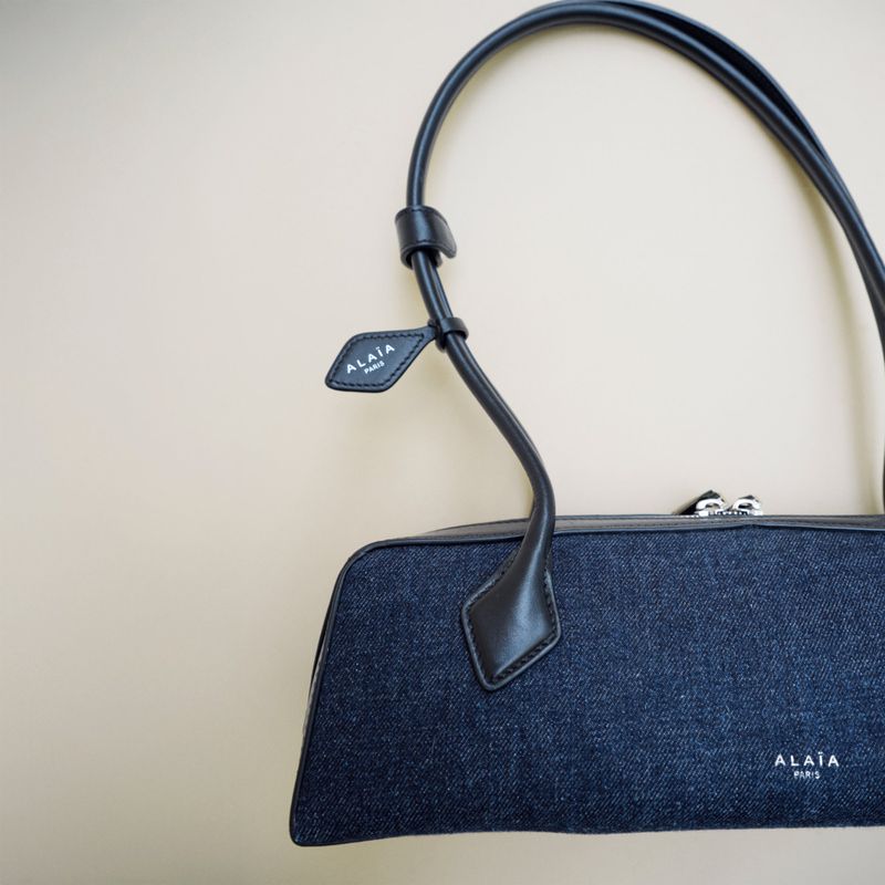 Alaïa Paris handbag featuring a blue denim exterior with black leather trim and handles on a beige background in a studio shot.