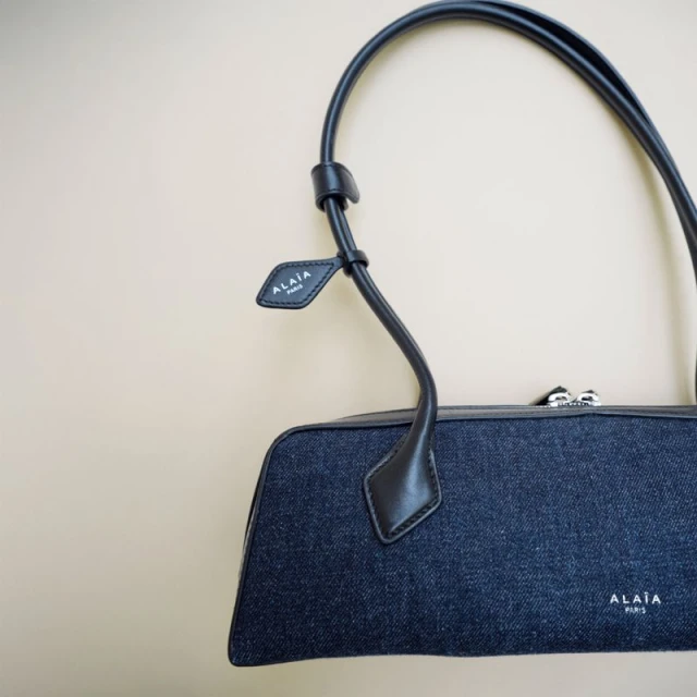 Alaïa Paris handbag featuring a blue denim exterior with black leather trim and handles on a beige background in a studio shot.