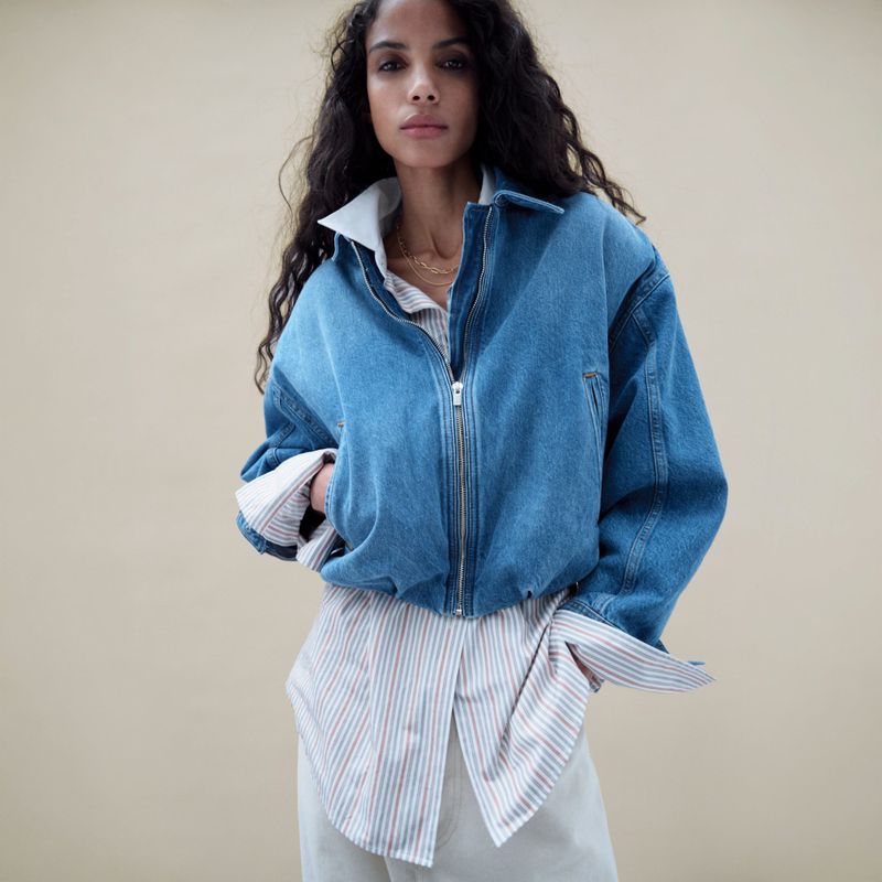 Adult woman wearing denim jacket and striped shirt posing in front of neutral backdrop. Fashion shot with natural light and relaxed pose.