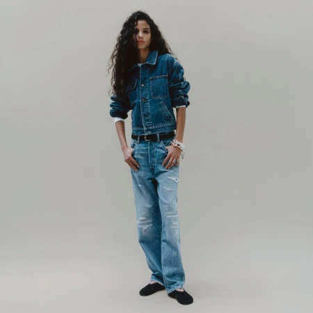 Model with long dark wavy hair wearing a denim jacket and blue jeans against a neutral grey studio backdrop.