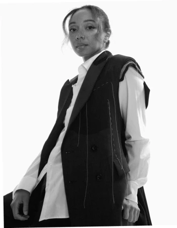 Black and white portrait of a stylish African American woman in a vest and white shirt, posing against a bright white background.