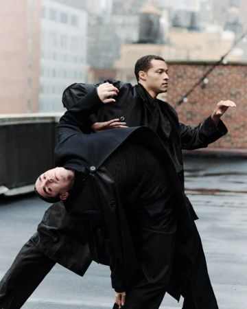 Two adult males perform a modern dance on a rooftop in black coats. An urban environment with soft, neutral light in the background.
