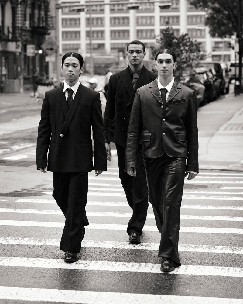 Three men in suits walk across a crosswalk in a black and white outdoor urban setting, portraying a classic and stylish group.