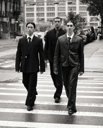 Three men in suits walk across a crosswalk in a black and white outdoor urban setting, portraying a classic and stylish group.