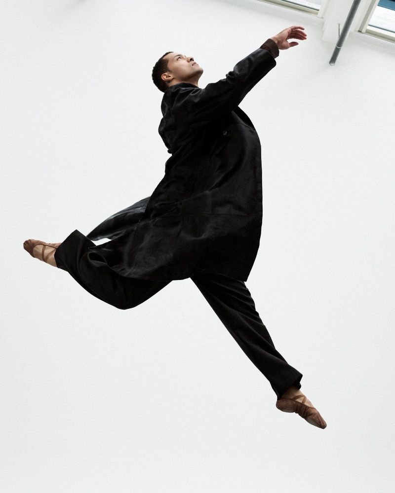 A man leaps in the air, wearing a black coat and pants with ballet shoes against a white background, captured from a low angle.