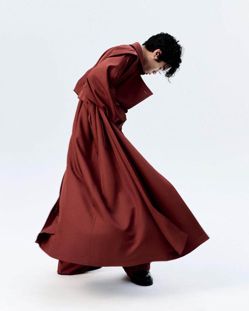 An adult male fashion model poses in a reddish-brown overcoat and dress pants in a bright studio. The model looks down and appears to be dancing.