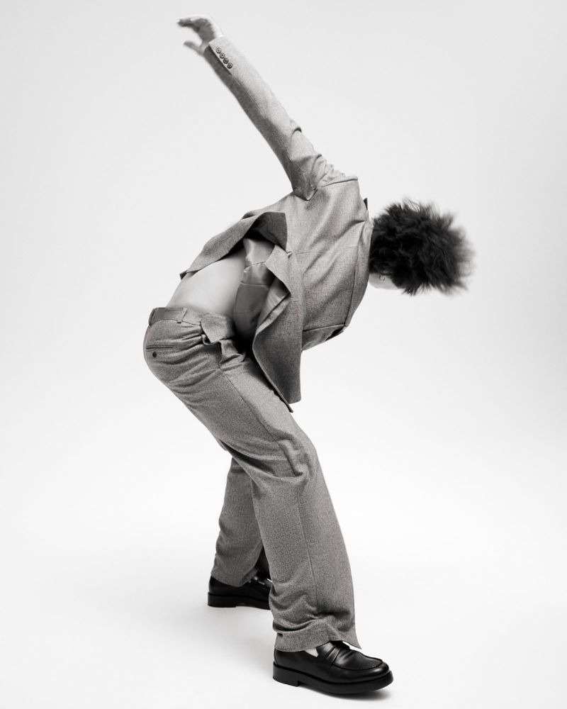 A person with curly hair is captured in a studio shot, wearing a suit with an open shirt, bent over with an arm extended overhead.