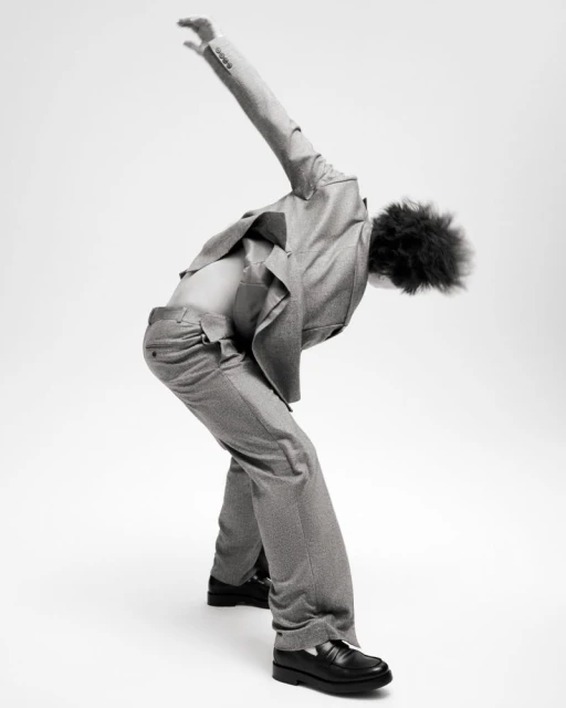 A person with curly hair is captured in a studio shot, wearing a suit with an open shirt, bent over with an arm extended overhead.