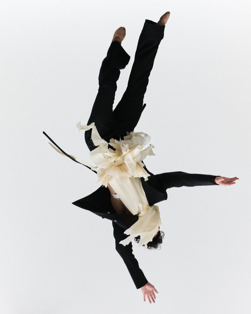 Person in mid-air wearing a black suit and ballet shoes, performing a dance move against a white background, captured in an action shot.