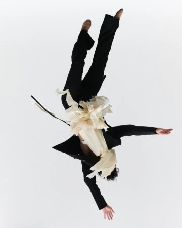Person in mid-air wearing a black suit and ballet shoes, performing a dance move against a white background, captured in an action shot.