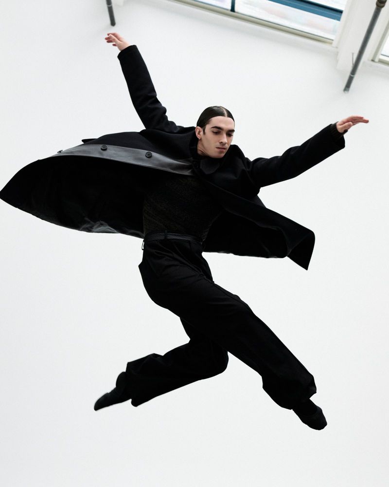 Stylish man in black suit and overcoat leaps mid-air in a graceful pose against a bright white background with window lights above.