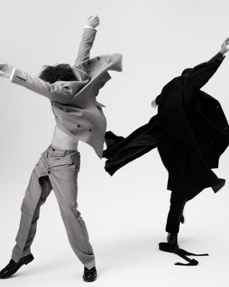 Two figures strike dynamic poses against a white background, one in an open grey suit, the other in a long black coat, capturing movement.
