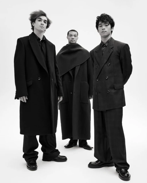 Three diverse young men in dark, fashionable coats and outfits stand in a minimalist studio against a white backdrop, exuding modern style.