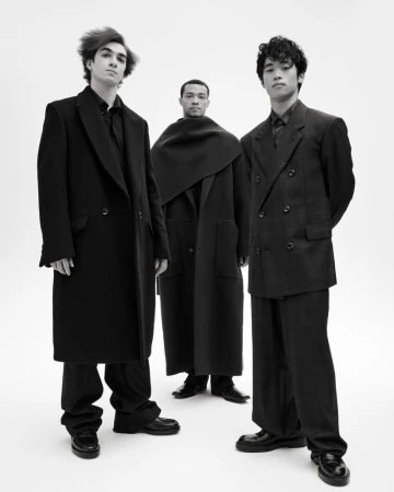 Three diverse young men in dark, fashionable coats and outfits stand in a minimalist studio against a white backdrop, exuding modern style.