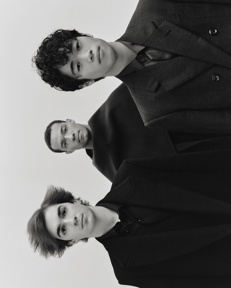 Black and white portrait of three young adult men standing close together in a photo studio wearing dark colored suit jackets.