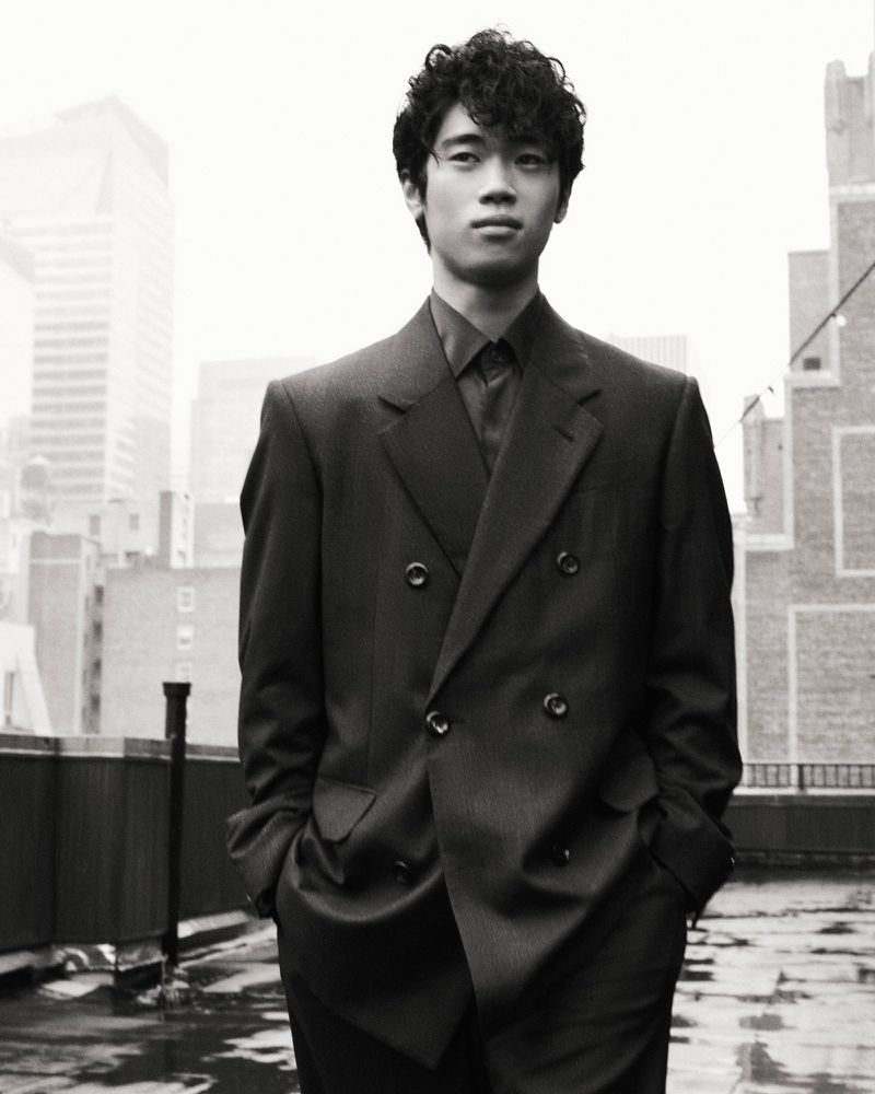 Black and white portrait of a curly-haired, Asian man in a suit, standing on a rooftop, with a city skyline in the background.