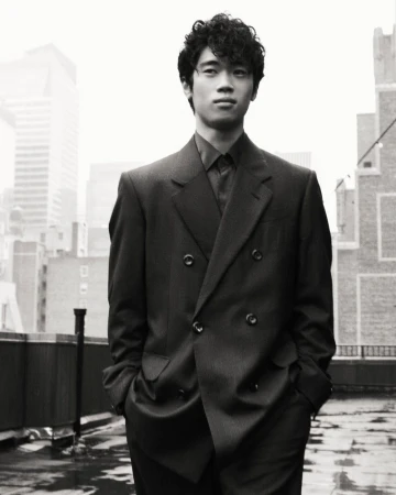 Black and white portrait of a curly-haired, Asian man in a suit, standing on a rooftop, with a city skyline in the background.