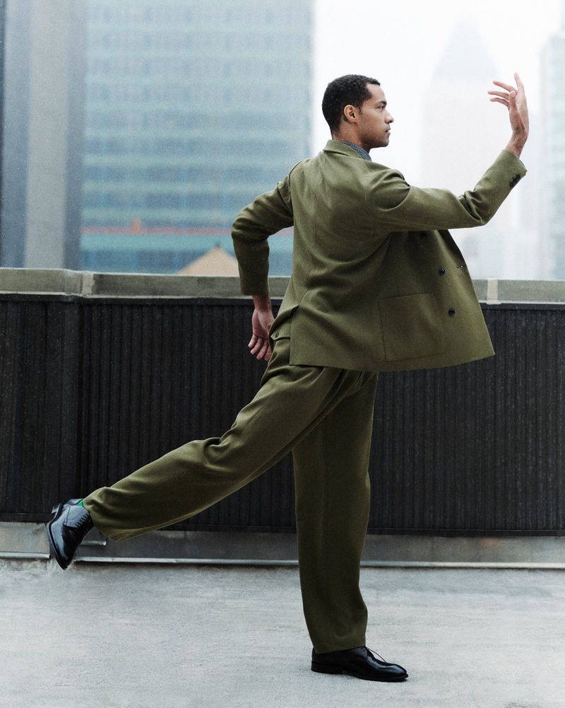 Man in green suit strikes ballet-inspired pose on rooftop. Modern skyscrapers in soft focus create an urban backdrop.
