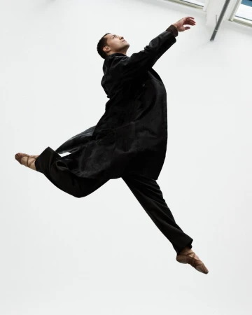 Dancer in a long dark coat and ballet shoes leaping mid-air against a bright white background