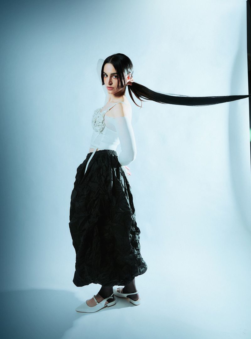 A full-length portrait of a woman in a white top and black skirt, in a photo studio with a light blue background and long ponytail.