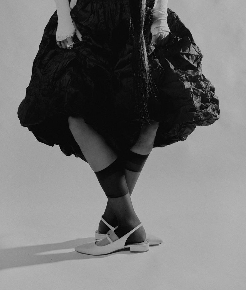 Fashion shot of person wearing black dress, white gloves, sheer black socks, and white shoes against a plain gray backdrop in studio lighting.