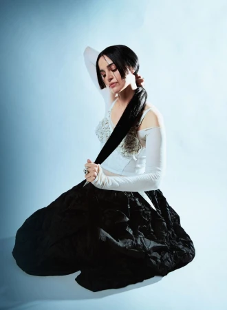 Portrait of a young adult woman with dark hair in a braid, kneeling in a white top and black skirt against a light blue backdrop.