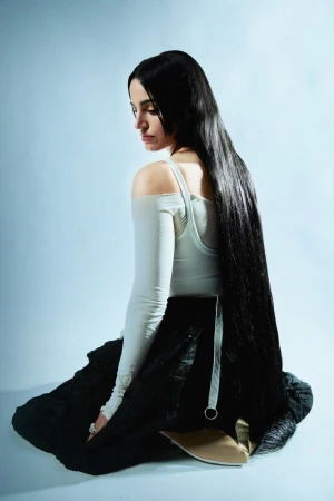 Brunette woman with long hair in a long sleeve top and skirt sitting in a light blue studio with pale neutral lighting. 
