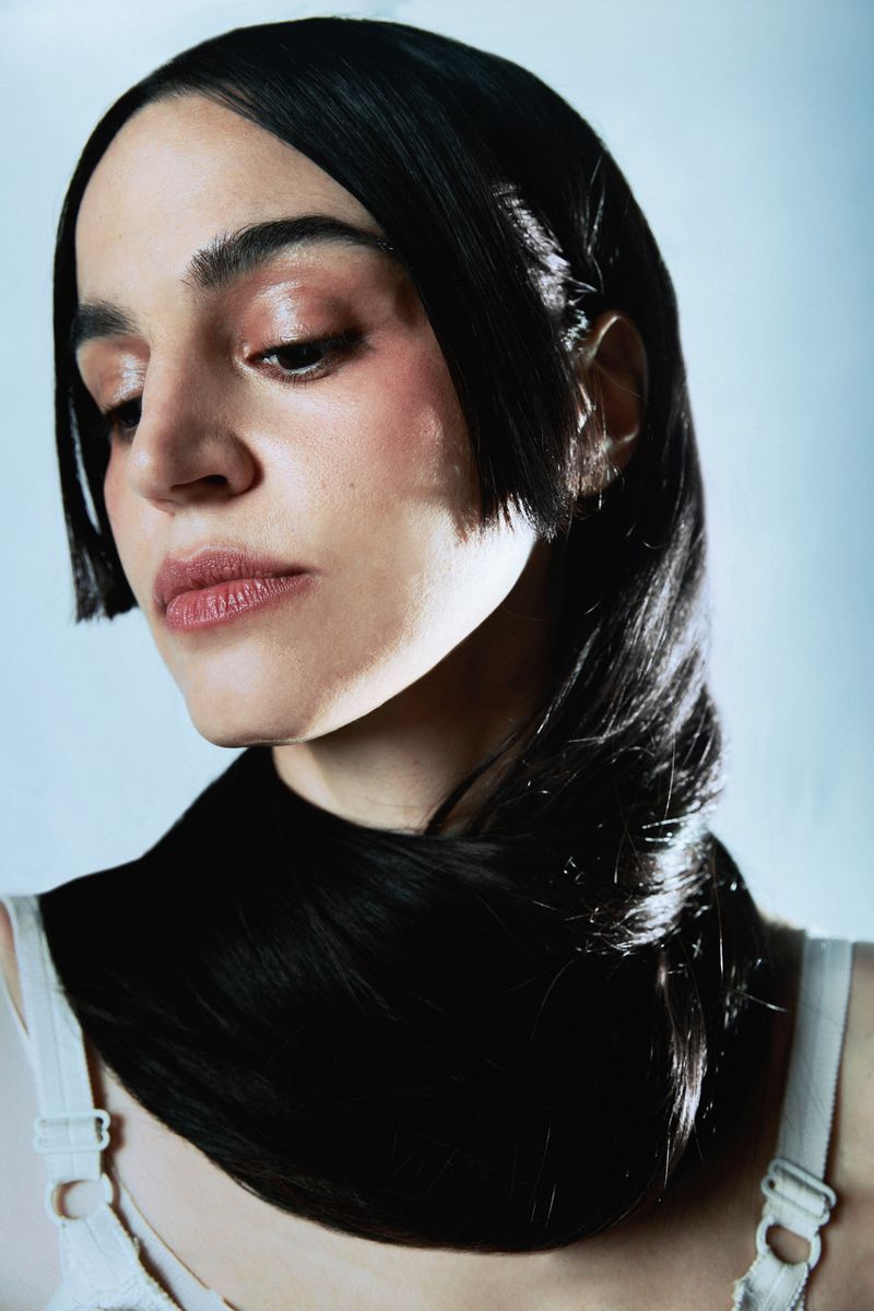 Close-up portrait of a woman with black hair styled around her neck, posing against a light blue background in a photo studio setting.