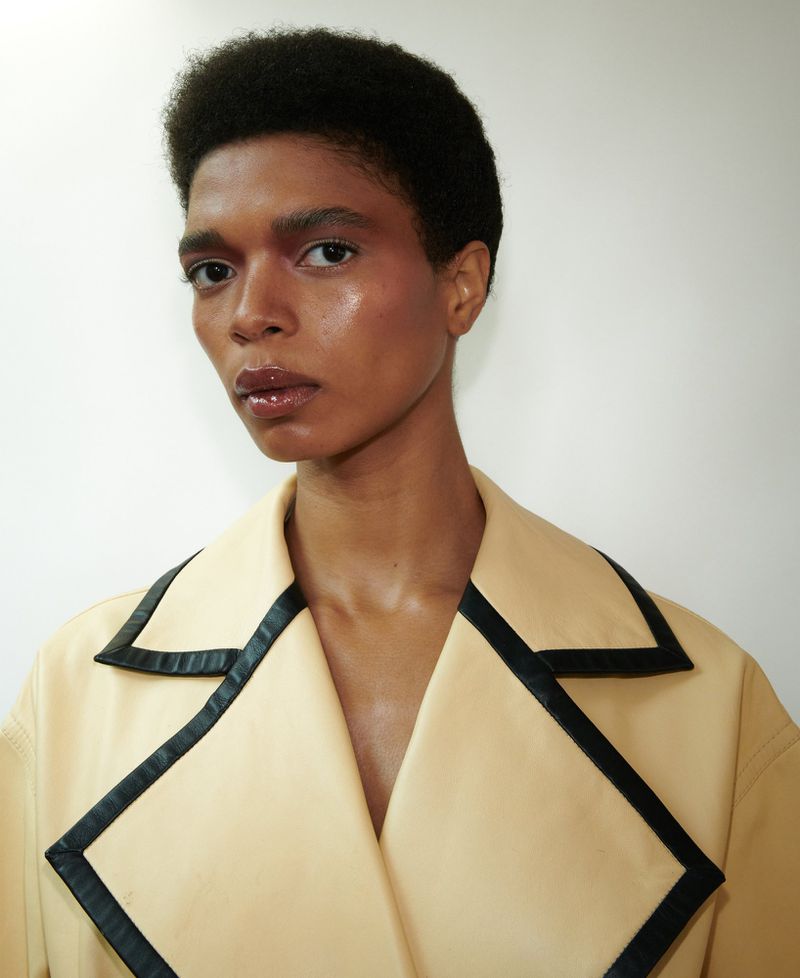 Portrait of an adult female with afro hairstyle and radiant makeup, wearing a light tan coat with a black trim in a studio setting.