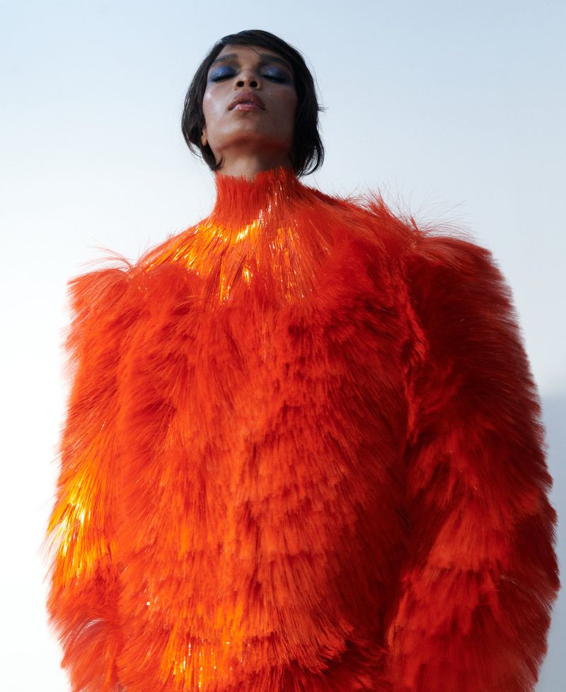 Woman in an orange textured dress poses against a white background with her eyes closed, showcasing fashion and beauty.