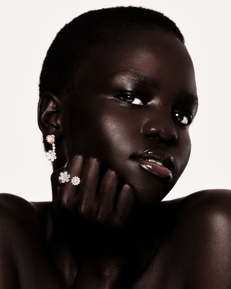 Close-up portrait of a dark-skinned woman with glossy lips, wearing diamond floral jewelry, against a clean white backdrop.
