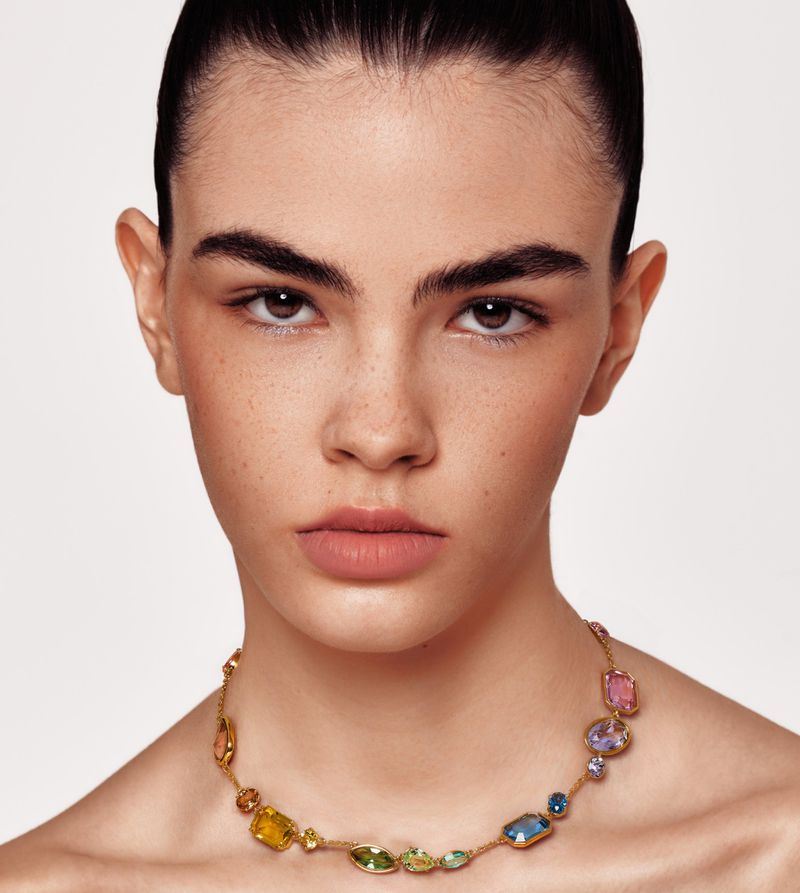 Close-up portrait of an adult woman with brown hair and fair skin wearing a gold chain necklace with colorful gemstone pendants.