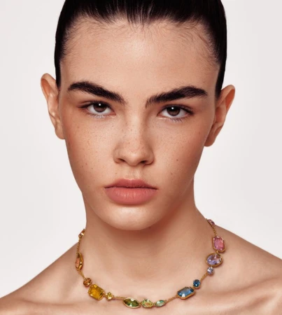 Close-up portrait of an adult woman with brown hair and fair skin wearing a gold chain necklace with colorful gemstone pendants.